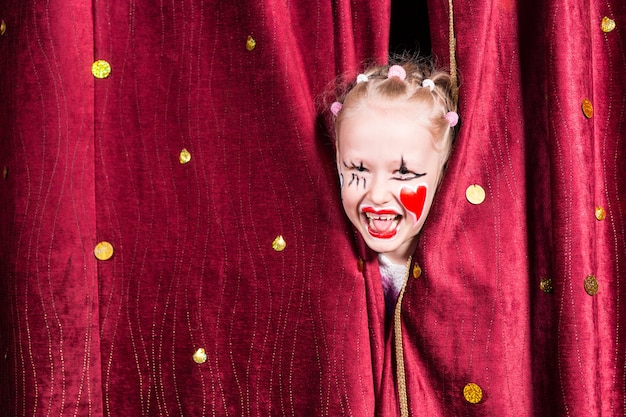 Pretty excited little blond girl on stage waiting to make her entrance on stage during a performance peeking out between the curtains and laughing