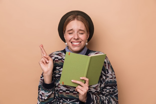 pretty excited woman in earrings holding diary and fingers crossed for good luck isolated over beige wall