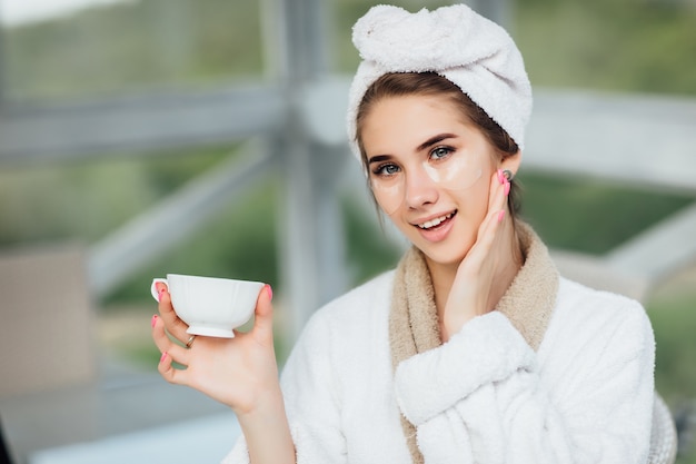 Pretty face. Attractive, smiling girl in white robe, sitting at hotel terrace and holding cup of coffee or tea. Make up concept.