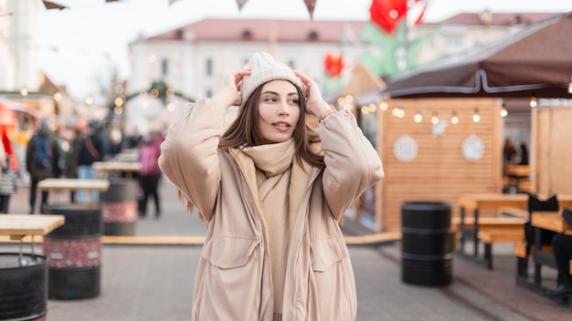 Pretty fashionable girl in stylish winter outerwear with a vintage knitted hat and beige jacket with a scarf walk in the city at the fair