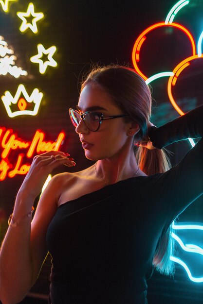 Pretty fashionable girl with glasses in a black stylish dress is fixing her hair on a dark background with colored neon signs light Night cool female portrait of beauty woman