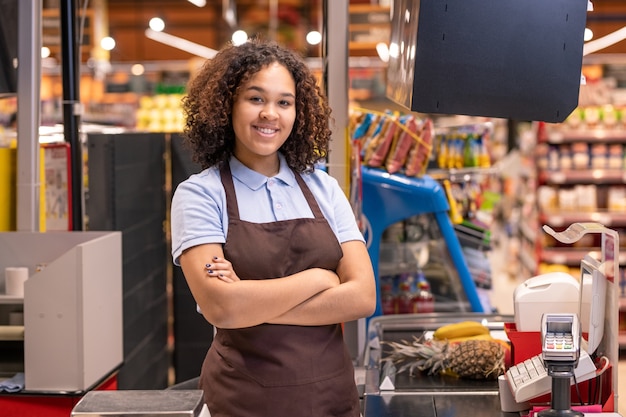 Pretty female in apron standing by cashbox in supermarket and crossing arms by chest on wall of shelves with food products