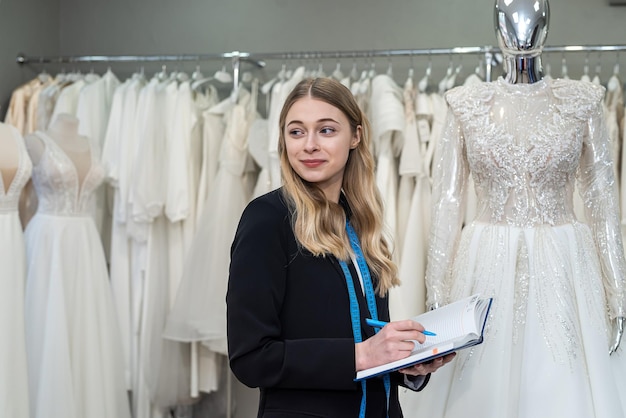 Pretty female designer or seller making notes near dress in wedding fashion store