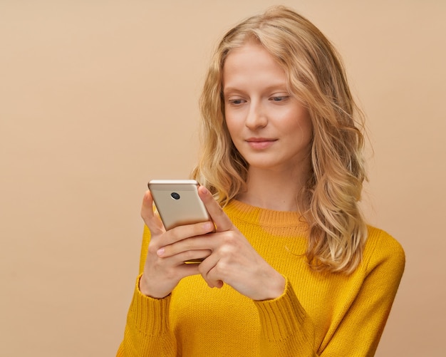 Pretty female looking social media on mobile phone standing near beige