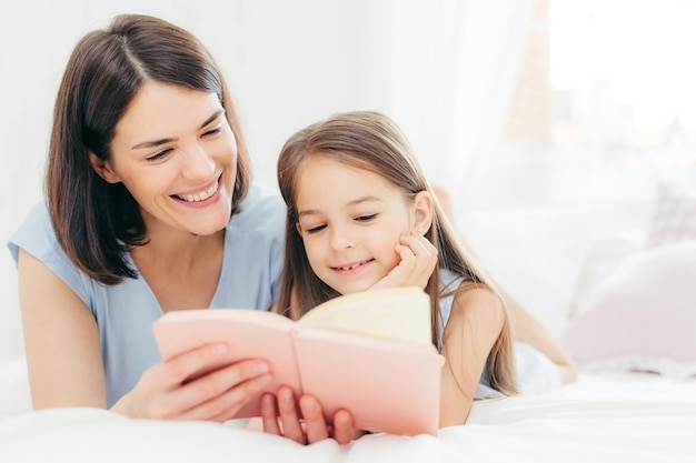 Pretty female mother and her small daughter read interesting book in bedroom lie on comfortabled bed during morning Lovely mum reads fairty tale for her little female kid who is very curious