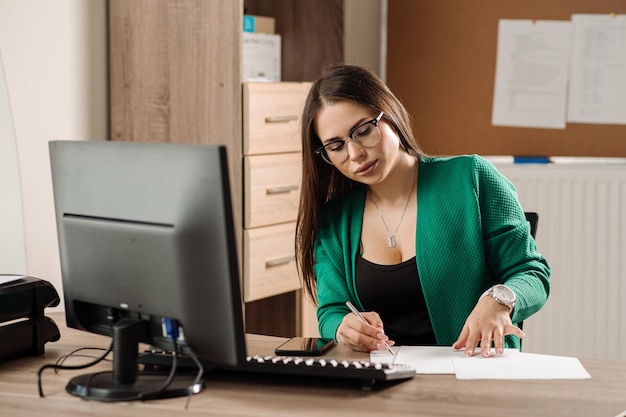 Pretty female office worker dressed in green jacket is sitting and working by the computer on her working place