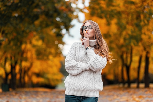 Pretty funny young woman with beautiful smile in glasses in a knitted sweater with a hairstyle is posing outdoors in the forest on a background of trees with orange-yellow leaves. Joyful hipster girl.