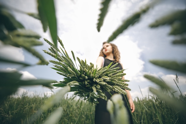 Pretty girl in black dress standing against cloudy sky with wheat bouquet in hands