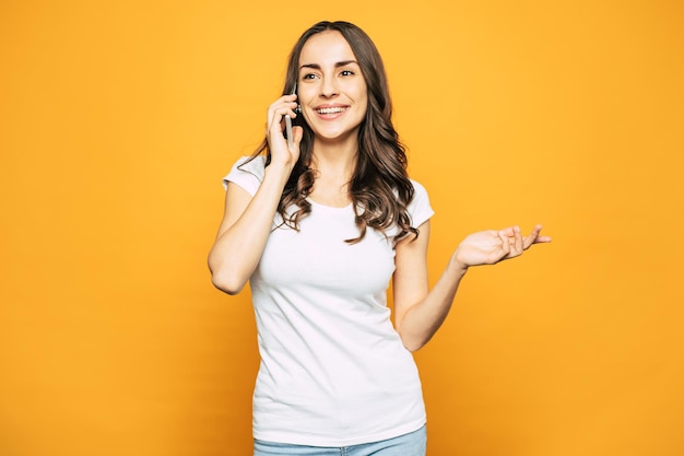 Pretty girl in casual outfit of T-shirt and denim jeans is talking to her boyfriend by mobile phone next to spice orange wall