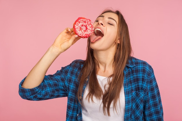 Pretty girl in checkered shirt standing with eyes closed and licking sweet donut dreaming of biting tasty doughnut struggling temptation to eat sugary confectionery indoor studio shot isolated