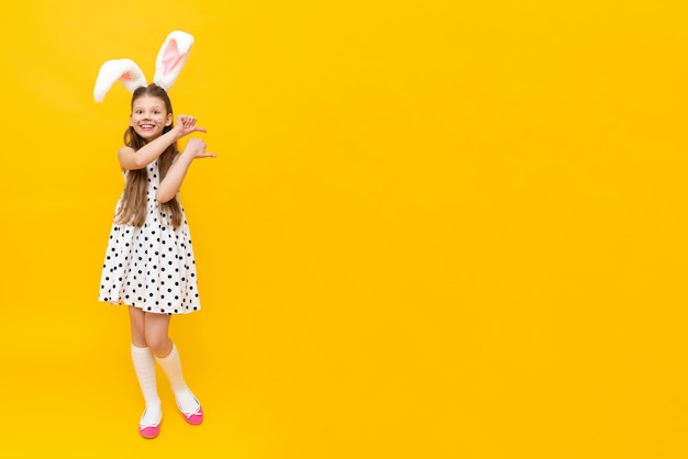 A pretty girl in fancy Easter bunny ears and painted mustache on a yellow isolated background shows your advertisement the spring Easter holiday