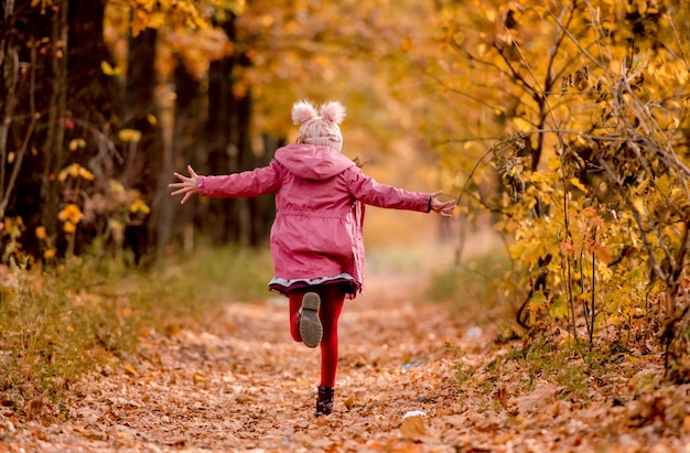 Pretty girl kid running at road with yellow leaves trees at autumn park. Beautiful portrait of child outdoors at nature