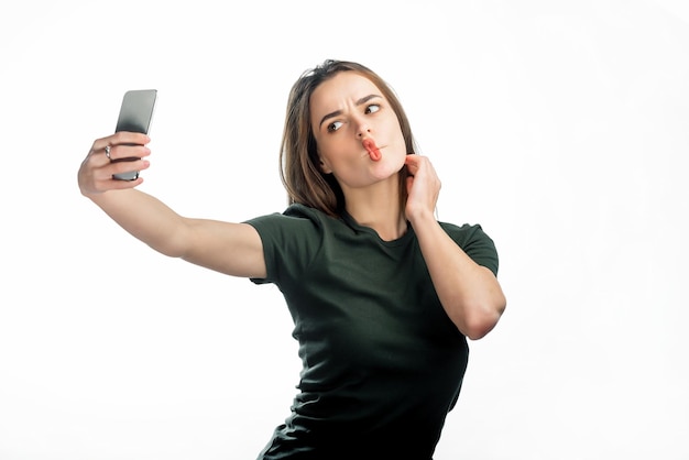 Pretty girl makes her lips duck and take a self portrait with her smart phone Cute lovely young woman making duck face and taking selfie with mobile phone over white background