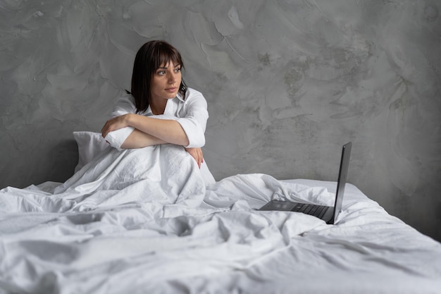 Pretty girl in the morning in the bedroom with a laptop looks out the window works remotely as a freelancer Relax and enjoy the weekend Notebook for coworking in the bedroom working house