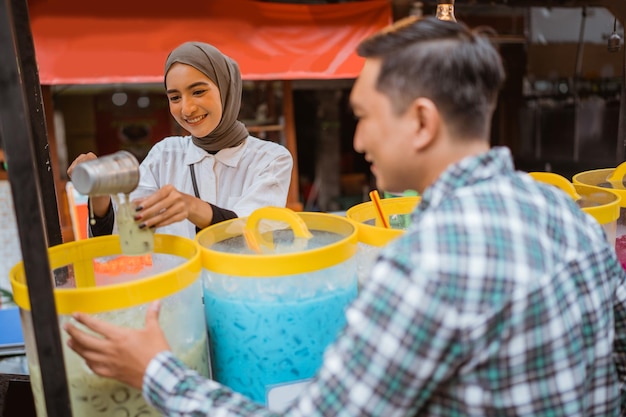 A pretty girl in a veil sells es campur using a scoop to get coconut milk from a jar on a cart