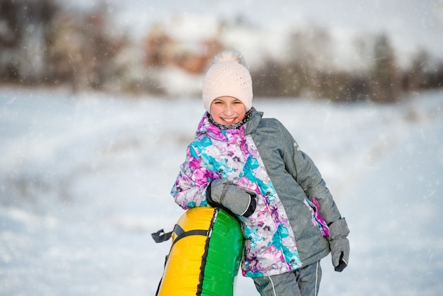 Pretty girl in warm clothes with inflatable snow sled standing on downhill at winter day