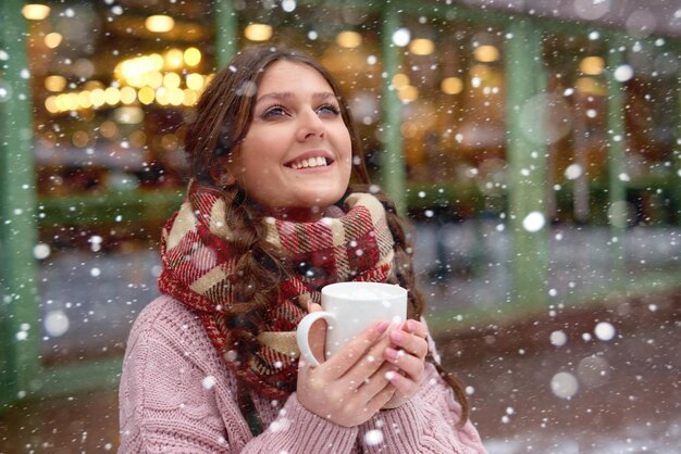 Pretty girl watching snow fall and drinking coffee or hot chocolate with marshmallows