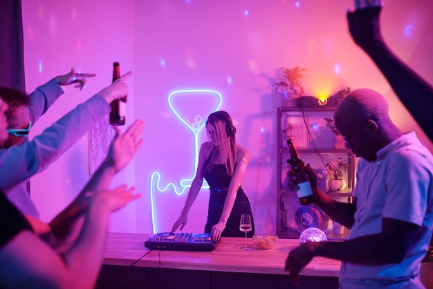 Pretty girl with long blond hair standing by counter with turntables while mixing music for disco dancing and enjoying at home party with friends