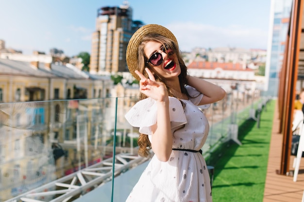 Pretty girl with long hair in sunglasses is posing to the camera on the terrace. She wears a white dress with bare shoulders, red lipstick and hat . She is smiling to the camera.