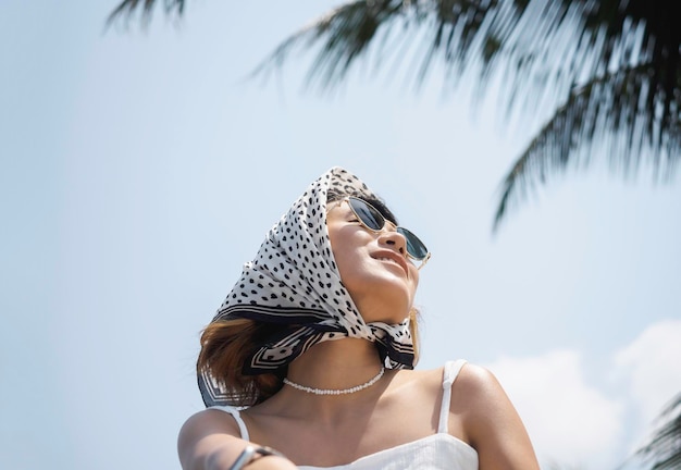 Pretty happy Asian woman portrait in casual white shirt wearing sunglasses and hair scarf enjoy with sunshine at the beach under the coconut palm trees and blue sky background in summer