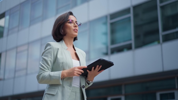 Pretty happy caucasian young woman in glasses having video chat on tablet device outdoor at business building