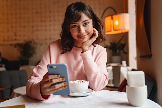 a pretty happy smiling brunette young woman indoors in cafe drinking coffee using mobile phone.