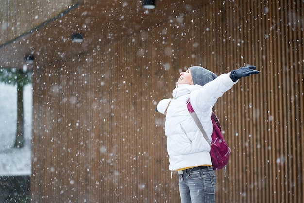 Pretty happy smiling woman with pink backpack wearing white jacket and grey jeans smiling and enjoying first snow with rised hands