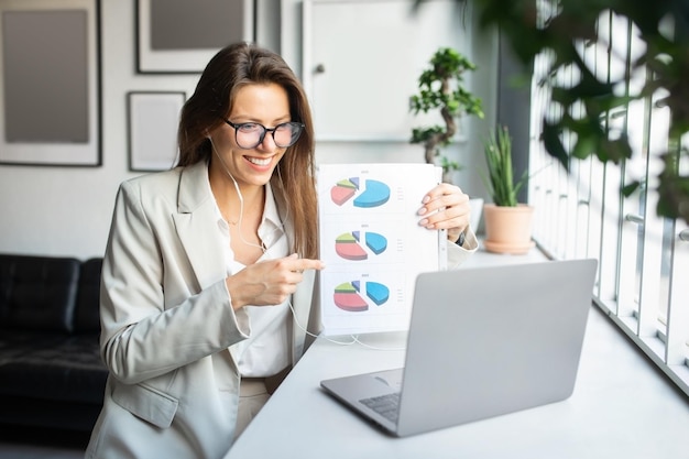 Pretty happy woman business consultant having online meeting via laptop showing paper with chart and