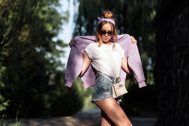 Pretty hipster girl with sunglasses and a bandana in denim shorts, a white T-shirt and a purple sweatshirt with a handbag walks outdoors