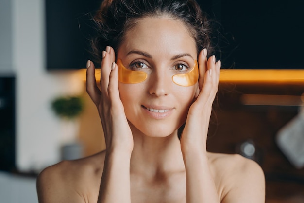 Pretty hispanic girl with naked shoulders moisturizes under eyes skin by patches Antiaging skincare