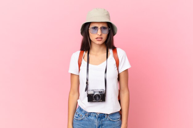 Pretty hispanic tourist feeling puzzled and confused with a photo camera and a hat