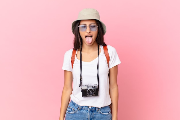 Pretty hispanic tourist with cheerful and rebellious attitude, joking and sticking tongue out with a photo camera and a hat