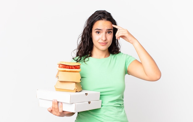 Pretty hispanic woman feeling confused and puzzled, showing you are insane and holding take away fast food boxes