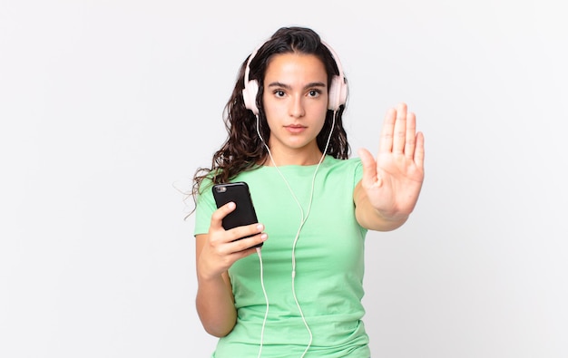 Pretty hispanic woman looking serious showing open palm making stop gesture with headphones and a smartphone