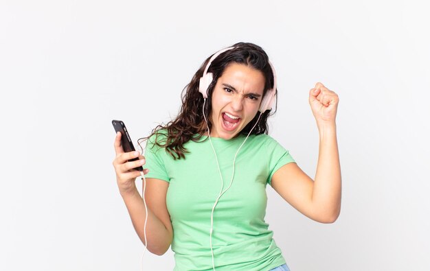 Photo pretty hispanic woman shouting aggressively with an angry expression with headphones and a smartphone