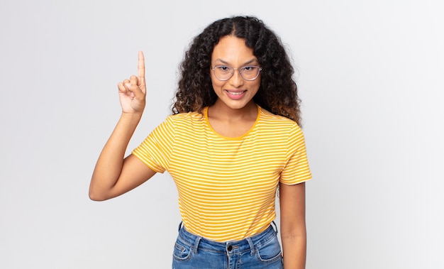 Pretty hispanic woman smiling cheerfully and happily, pointing upwards with one hand to copy space