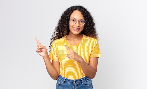 pretty hispanic woman smiling happily and pointing to side and upwards with both hands showing object in copy space