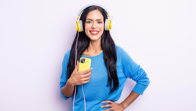 Pretty hispanic woman smiling happily with a hand on hip and confident. smartphone and headphones concept