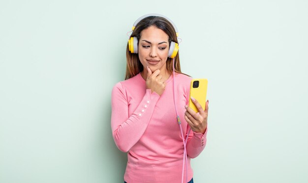 Pretty hispanic woman smiling with a happy, confident expression with hand on chin. smartphone and headphones concept