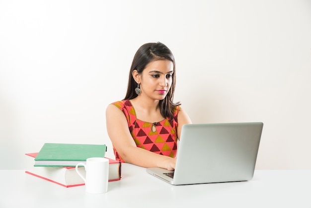 Pretty Indian Asian Girl studying on laptop computer with pile of books on table, over white background