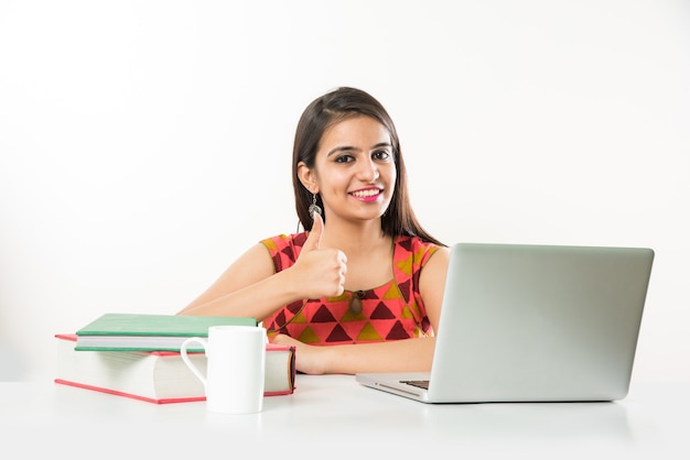 Pretty Indian Asian Girl studying on laptop computer with pile of books on table, over white background