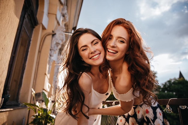 Pretty ladies in good humor pose on terrace Curly girls in summer light clothes are photographed against clear blue sky