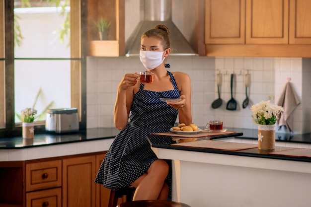 Pretty lady in a medical mask sits in the kitchen and drinks  herbal tea. Quarantine. Stay at home. Self-isolation