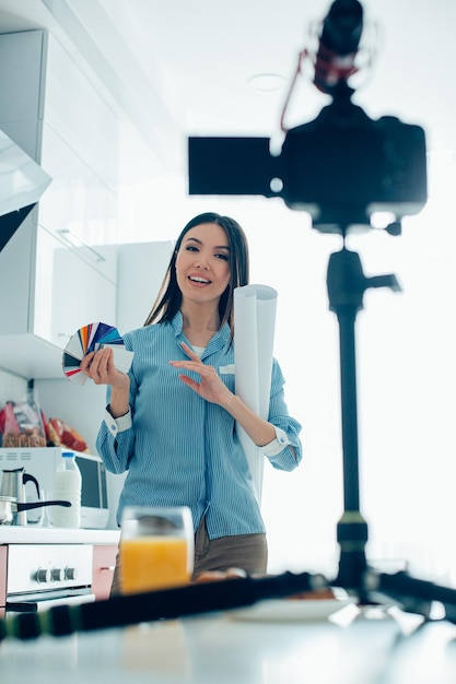 Pretty lady standing with a paper roll and color swatches while talking on the camera in her kitchen