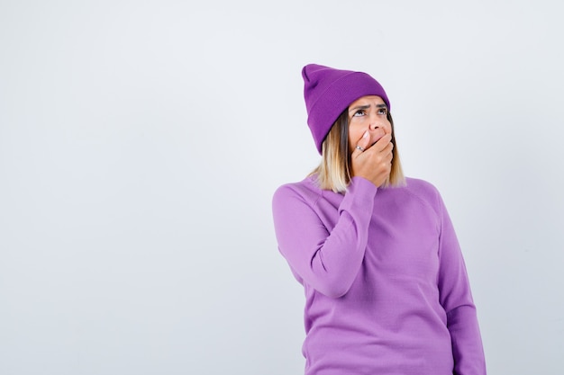 Pretty lady in sweater, beanie with hand on open mouth, looking up and looking surprised , front view.