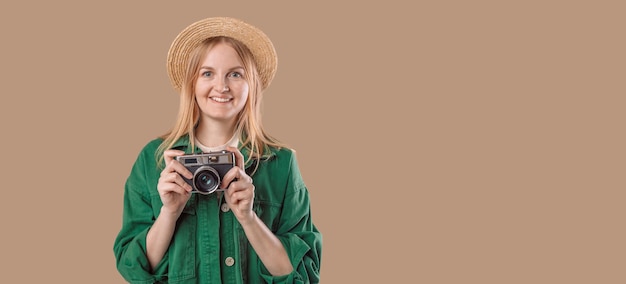 Pretty laughing woman in summer hat and green jacket holding vintage camera and posing over background in studio