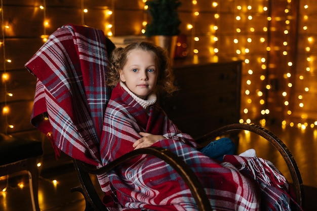 Pretty little blonde curly child girl wrapped in plaid looking at camera sitting in rocking chair at