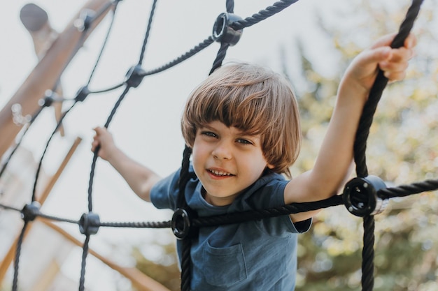 Pretty little brunette boy in a blue T-shirt is climbing a rope web. child is engaged in outdoor sports. healthy active kid. lifestyle. space for text. High quality photo