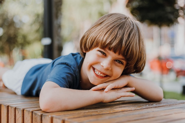 Pretty little brunette boy in a blue T-shirt and white shorts is lying on a wooden bench in park. child sunbathes and takes an air bath outdoors. concept of health. space for text. High quality photo