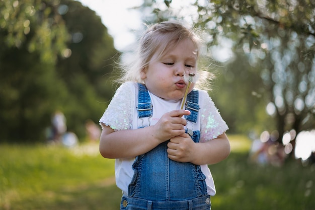 Pretty little girl blowing off dandelion seeds on sunset in summer park Image with selective focus